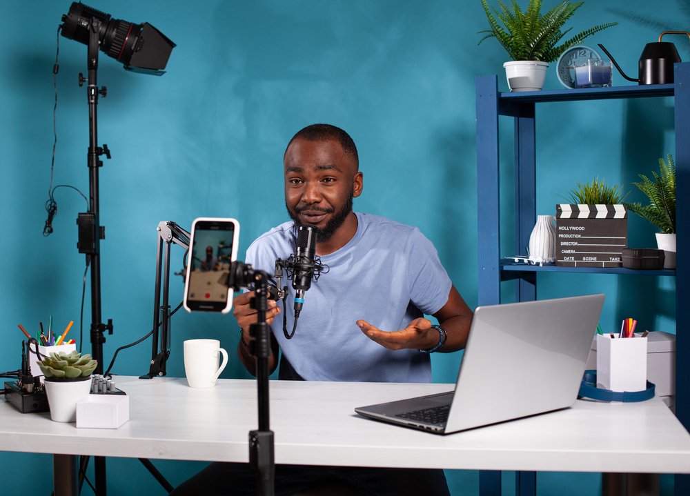 How do brand deals work -- Influencer sitting down at desk with laptop in vlogging studio smiling in front of recording smartphone holding microphone. Content creator interacting with fans looking at live video podcast setup.